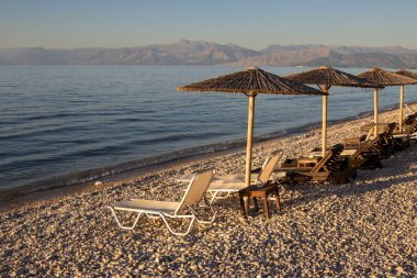 İyon Denizi 'nin çakıl taşı plajı. Boş şezlonglar ve tahta güneş şemsiyeleri. Arkadaki Arnavut dağları. Mavi gökyüzü. Gün batımına. Acharavi, Korfu (Korfu), Yunanistan.