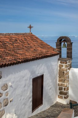 Beyaz cepheli, çapraz ve turuncu kiremitleri olan eski küçük bir kilise. Tek zilli Felfry kulesi. Taş detaylar. Atlantik Okyanusu ve açık beyaz bulutlu mavi gökyüzü. San Juan de la Rambla, Tenerife, İspanya.