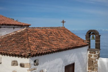 Beyaz cepheli, çapraz ve turuncu kiremitleri olan eski küçük bir kilise. Tek zilli Felfry kulesi. Taş detaylar. Atlantik Okyanusu ve açık beyaz bulutlu mavi gökyüzü. San Juan de la Rambla, Tenerife, İspanya.