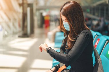Tıbbi koruyucu yüz maskesi takan Asyalı kadın havaalanı, sağlık ve enfeksiyon konseptindeki terminal dinlenme odasında saatlerini kontrol etmek için kol saatine bakıyor..