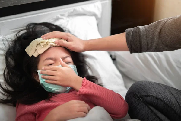 アジアの母親は 病気の少女額に高熱を減らすためのクールなタオルを取ります 女の子は医療保護顔マスクと咳を身に着けています 選択的な焦点 健康と感染の概念 — ストック写真