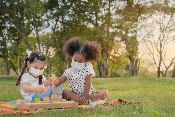 幸せな陽気な民族の女の子が一緒に屋外公園で木製のブロックパズルをプレイ 彼らはウイルスからそれらを保護するためにマスクを着用 関係の小さな子供 多様な民族の概念 — ストック写真