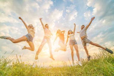 Mutlu arkadaşlar güneşli arka planda çimlerin üzerinde zıplıyorlar.