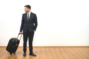 Beyaz duvar arkasında duran bavullu mutlu iş adamı.