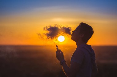 Adam günbatımında arka planda elektronik sigara içiyor.
