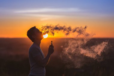 Adam arka planda günbatımında elektrikli sigara içiyor.