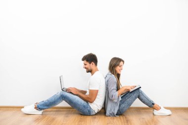 Gülümseyen çift boş duvarın yanında dizüstü bilgisayar ve tabletle oturuyor.