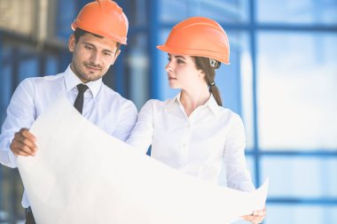 İki mühendis binanın arka planındaki proje planına bakıyor.