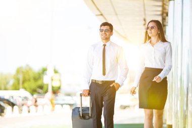Güneş gözlüklü iş adamı ve kadın bir bavulla yürüyorlar.