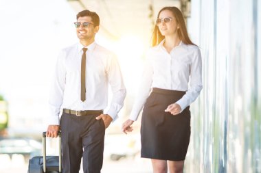 Güneş gözlüklü iş adamı ve kadın bir bavulla yürüyorlar.