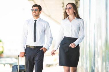 Güneş gözlüklü mutlu iş adamı ve kadın bir bavulla yürür.