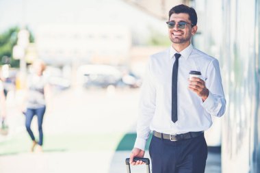 Güneş gözlüklü işadamı kahve fincanını tutar ve bir bavulla yürür.