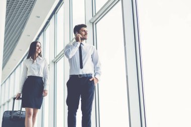 Kadın ve erkek panoramik pencerelerin yanında bir bavul ve telefonla yürüyorlar.