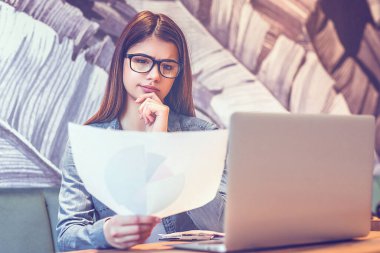 Masada dizüstü bilgisayarın yanında gözlüklü bir kadın gazete tutuyor.
