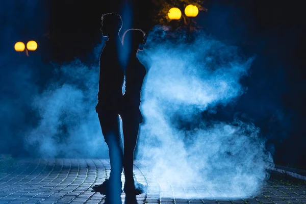 Adam ve kadın, karanlık bir sokakta mavi bir duman arka planında dikiliyorlar. Gece vakti.