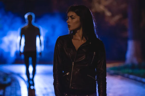 Kadın dumanlı bir adamın arka planında karanlık bir sokakta duruyor. Gece vakti.