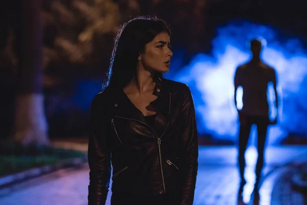 Kadın dumanlı bir adamın arka planında karanlık bir sokakta duruyor. Gece vakti.