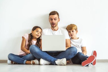Dizüstü bilgisayarı olan baba ve çocuklar yerde oturuyor.