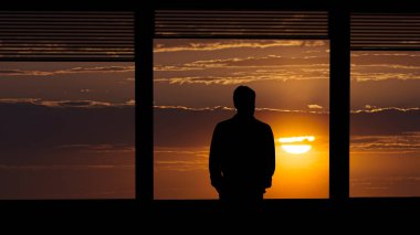 Günbatımı arka planında pencerenin yanında duran adam.