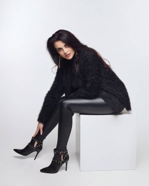 Uzun saçlı çekici esmer beyaz bir küpün üzerinde oturuyor. Stüdyoda beyaz arkaplanda fotoğraf çekimi