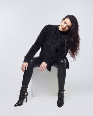 Uzun saçlı çekici esmer beyaz bir küpün üzerinde oturuyor. Stüdyoda beyaz arkaplanda fotoğraf çekimi