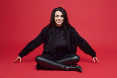 Çekici esmer, uzun saçlı, kırmızı arka planda stüdyoda oturuyor.