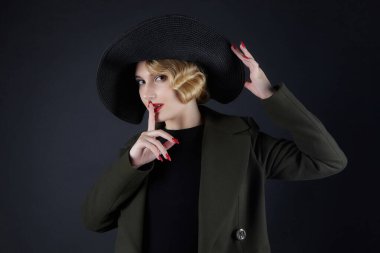 Siyah şapkalı, geçmişe dönük saçları olan çekici sarışın dudaklarına parmağını bastırmış.