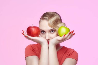 Geçmişe dönük saç stili ile çekici genç bir sarışın kırmızı ve yeşil bir elmayı yüzünün önünde tutuyor.