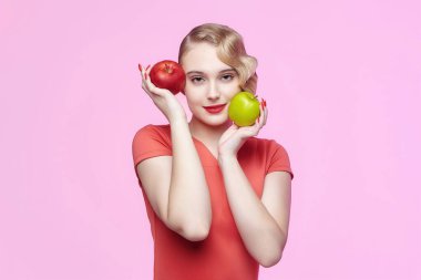 Geçmişe dönük saç stili ile çekici genç bir sarışın kırmızı ve yeşil bir elmayı yüzünün önünde tutuyor.