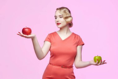Geçmişe dönük saç stili olan çekici genç bir sarışın elinde kırmızı ve yeşil bir elma tutuyor. Pembe arka planda fotoğraf çekimi