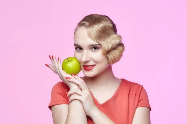 Eski moda saç stili olan çekici genç bir sarışın. Yüzünün yanında yeşil bir elma tutuyor.