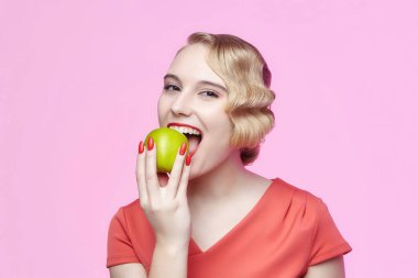 Geçmişe dönük saç stili olan çekici sarışın yeşil bir elmayı ısırmak üzere.