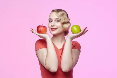 Geçmişe dönük saç stili ile çekici genç bir sarışın kırmızı ve yeşil bir elmayı yüzünün önünde tutuyor.