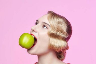 Eski saç stili olan çekici genç bir sarışın dişlerinde yeşil bir elma tutuyor.