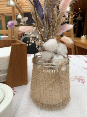 Masanın üstündeki vazoda beyaz bir masa örtüsüyle pamuk çiçek buketi. Kafe iç ahşap dizayn, kırsal tarzda. Bir buket kurumuş çiçek ve peçete tutacağı.