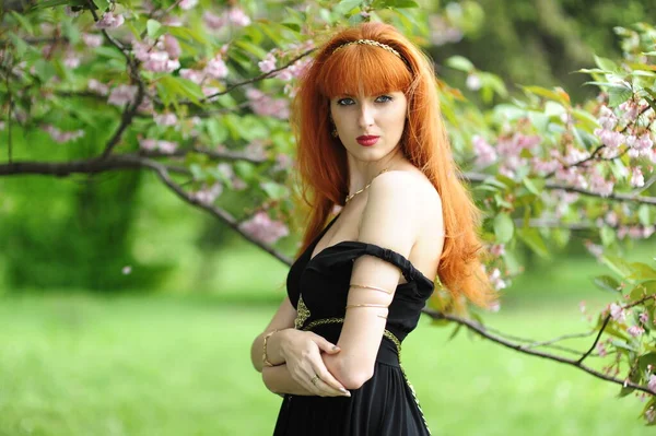 Jeune belle femme aux cheveux roux dans une robe longue noire dans un jardin de printemps sur un fond de sakura en fleurs — Photo