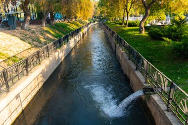 Park yolları ve yeşil ağaçlar arasında akan temiz suyu olan şehir kanalı. Demiryolu ve küçük su çıkışlı güneşli bir gün sahnesi, şehir sulama ve manzara tasarımı konsepti