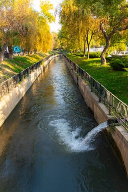 Park yolları ve yeşil ağaçlar arasında akan temiz suyu olan şehir kanalı. Demiryolu ve küçük su çıkışlı güneşli bir gün sahnesi, şehir sulama ve manzara tasarımı konsepti