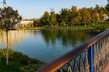 Sakin gölü olan bir park manzarası ve gün batımında suya yansıyan yeşil ağaçlar. Huzurlu kentsel doğa sahnesi, önünde dekoratif bir köprü var.