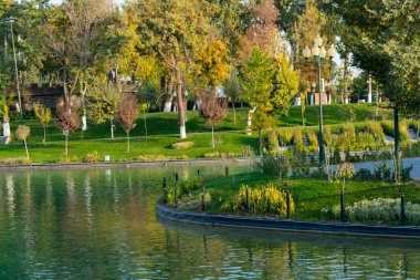 Sakin gölü olan bir park manzarası ve gün batımında suya yansıyan yeşil ağaçlar. Huzurlu kentsel doğa sahnesi, önünde dekoratif bir köprü var.