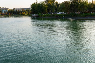 Sakin gölü olan bir park manzarası ve gün batımında suya yansıyan yeşil ağaçlar. Huzurlu kentsel doğa sahnesi, önünde dekoratif bir köprü var.