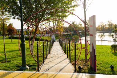 Ağaçlı yeşil park, dekoratif çitler ve güneşli bir öğleden sonra göl. Yürüyüş yolu ve sokak lambasıyla huzurlu kentsel dinlenme alanı