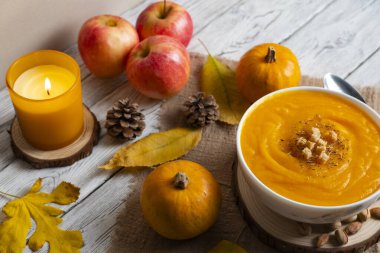 Sıcak sonbahar sahnesinde kremalı balkabağı çorbası, kızarmış ekmek, elma, balkabağı, kozalak ve mum ışığı. Sonbahar tarifleri, yemek blogları ve mevsimsel promosyonlar için mükemmel.