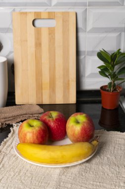 Kırmızı elmalı beyaz bir tabak ve bej bir altlık üzerinde olgun bir muz yanında da çıtır ekmek ve bir kupa. Sıcak mutfak kurulumu sağlıklı beslenme temaları için idealdir