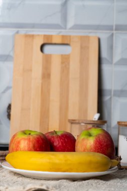 Kırmızı elmalı beyaz bir tabak ve bej bir altlık üzerinde olgun bir muz yanında da çıtır ekmek ve bir kupa. Sıcak mutfak kurulumu sağlıklı beslenme temaları için idealdir