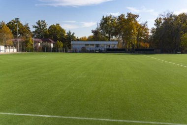 Güneşli bir günde yeşil suni çimleri olan boş bir futbol sahası. Sonbahar ağaçları ve açık gökyüzü olan bir spor sahası. Açık havada atletizm geçmişi