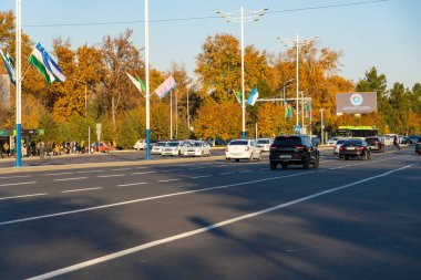 Taşkent, Özbekistan, 15 Kasım 2025: Sıcak akşam ışığında, parlak sonbahar ağaçlarıyla çevrili bir şehir caddesinde arabalar sürüyor. Şehir trafiği, mevsimsel atmosfer ve modern yol altyapısı