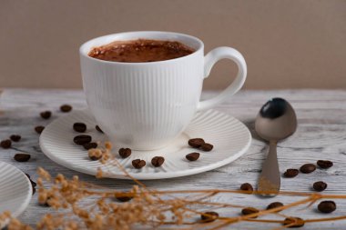 Bir fincan sıcak köpüklü kahve, beyaz bir çay tabağının üzerinde, kahve çekirdekleri de ahşap bir masanın üzerinde. Üst manzara, rahat sabah atmosferi, minimal yiyecek ve içecek kompozisyonu.