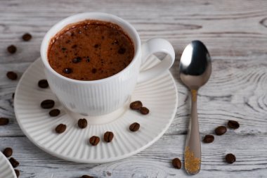 Bir fincan sıcak köpüklü kahve, beyaz bir çay tabağının üzerinde, kahve çekirdekleri de ahşap bir masanın üzerinde. Üst manzara, rahat sabah atmosferi, minimal yiyecek ve içecek kompozisyonu.