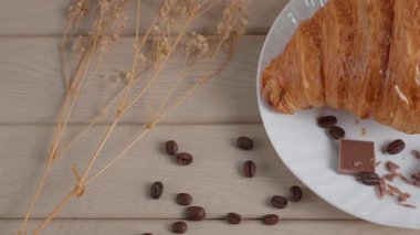 Kahve çekirdekleri ve kurumuş çiçeklerle beyaz bir tabakta çikolatalı kruvasanın sinematik yakın çekimi. Pürüzsüz kamera hareketi desen, fırın havası, kahvaltı veya kafe kavramını vurgular